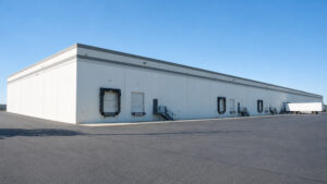 Long white industrial warehouse with loading docks and a parked semi-trailer on a wide asphalt lot under a clear blue sky, left-side perspective