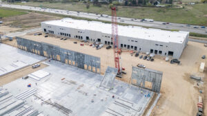 Aerial view of a large construction site with a red crane, partially built concrete walls, and a long white warehouse under construction near a highway.