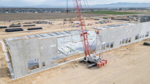 Construction site with a large concrete building under erection; a red crane lifts panels as workers guide them into place.