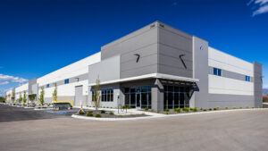 Modern industrial building with a gray facade, glass entry doors, and a bright blue sky; small landscaping and a scissor lift parked near the entrance.