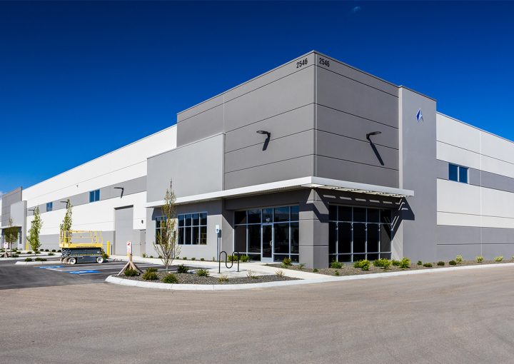 Modern industrial building with a gray facade, glass entry doors, and a bright blue sky; small landscaping and a scissor lift parked near the entrance.