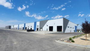 Exterior of a modern industrial warehouse with loading bays, blue and gray panels, and a wide paved yard under a clear blue sky.