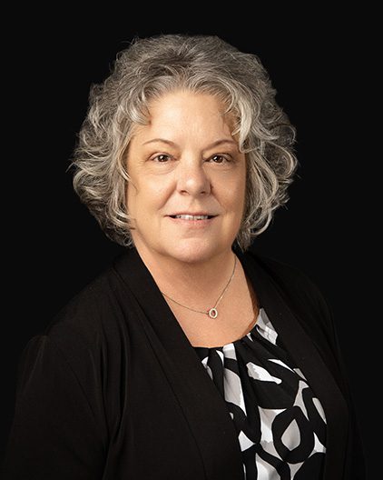 Portrait of a smiling, middle‑aged woman with short gray hair, wearing a black cardigan and a black‑and‑white blouse against a dark background.