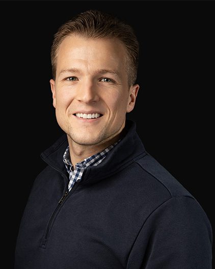 Portrait of a smiling man wearing a navy quarter-zip sweater against a black background, head and shoulders visible.