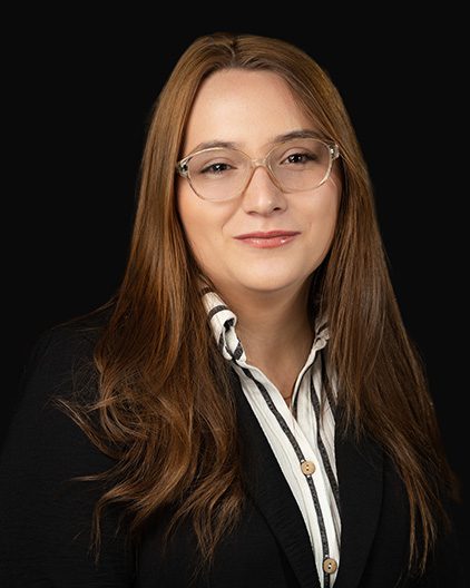 Professional headshot of a woman with long brown hair and glasses, wearing a black blazer and striped shirt against a dark background.
