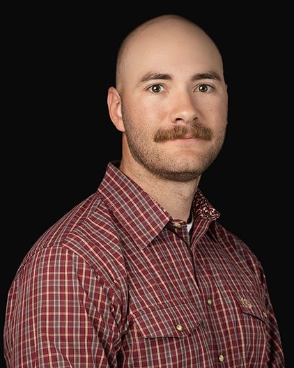 Portrait of a bald man with a mustache wearing a red plaid shirt against a black background.