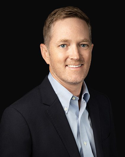 Professional headshot of a smiling man in a navy blazer and light blue shirt against a dark background.