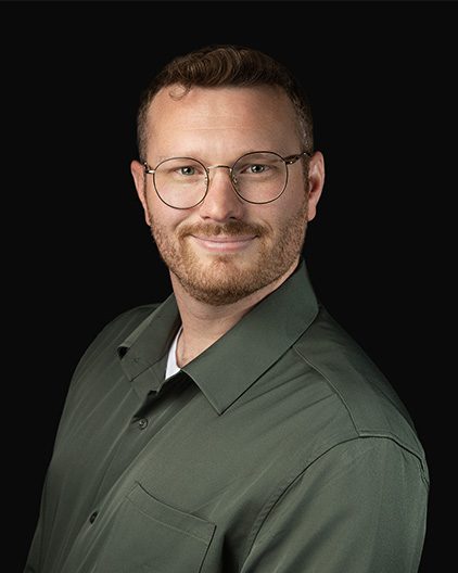 Head-and-shoulders portrait of a smiling man with short brown hair and round glasses, wearing an olive-green button-up shirt against a black background.
