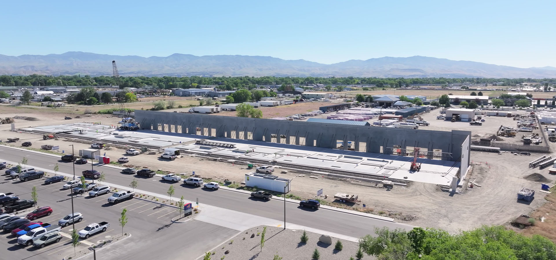 idaho industrial construction team | Adler Industrial Aerial view of a construction site with a long gray building under construction, heavy equipment, and a road with parked cars in the foreground, mountains in the distance.