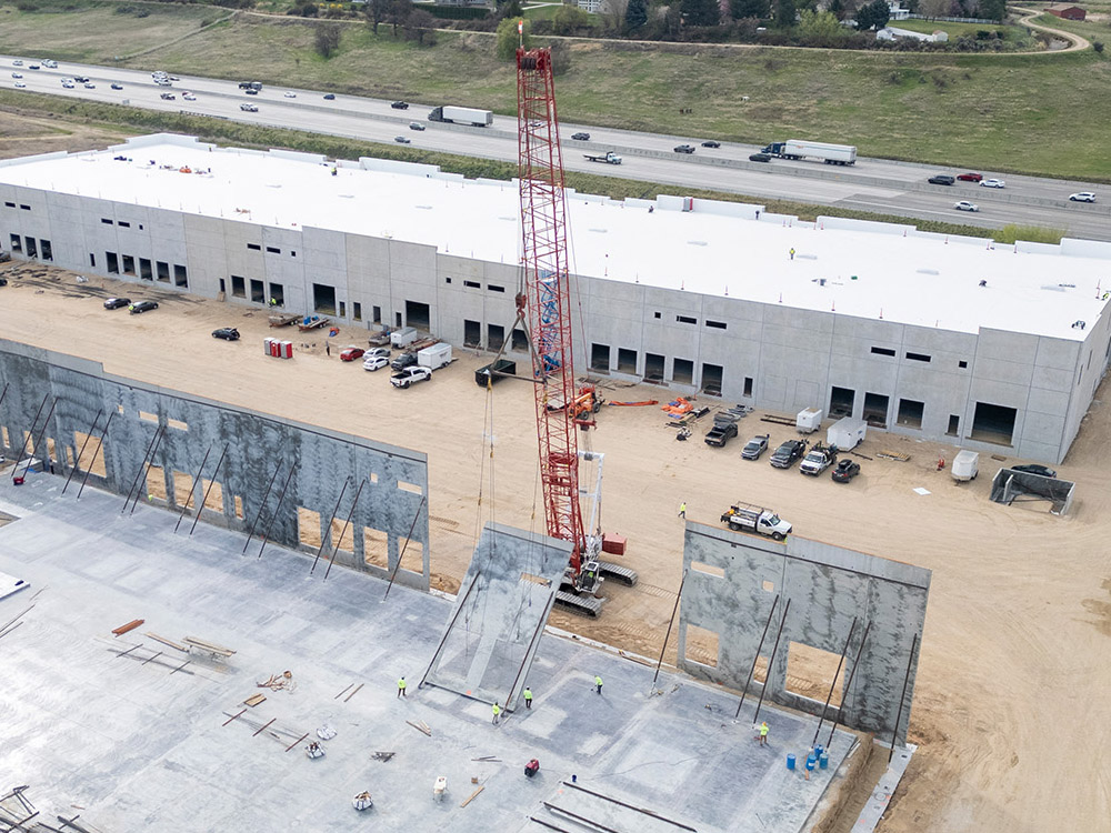 Aerial view of a large construction site with a red crane beside a long grey industrial building, workers and vehicles scattered around the dirt lot, and a highway nearby.